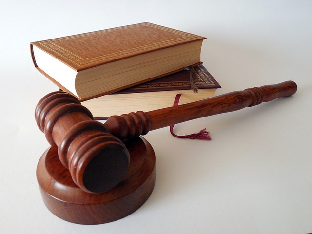 A judge's hammer rests on a resonance block. Both are made of dark brown wood, probably oak. Behind it lies a stack of two books. The upper book is bound in a light brown leather cover and the lower one in a slightly darker leather cover. A fabric bookmark hangs out of the lower book. All the items lie on a white background.