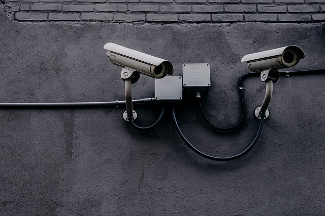 Two silver, elongated surveillance cameras hang on a wall, connected to two power boxes behind them via pipes and cables. The first camera is tilted slightly downwards to the right, the second camera is tilted slightly higher but also points to the right. Most of the wall is plastered with gray plaster. Only the upper edge of the picture shows gray bricks.